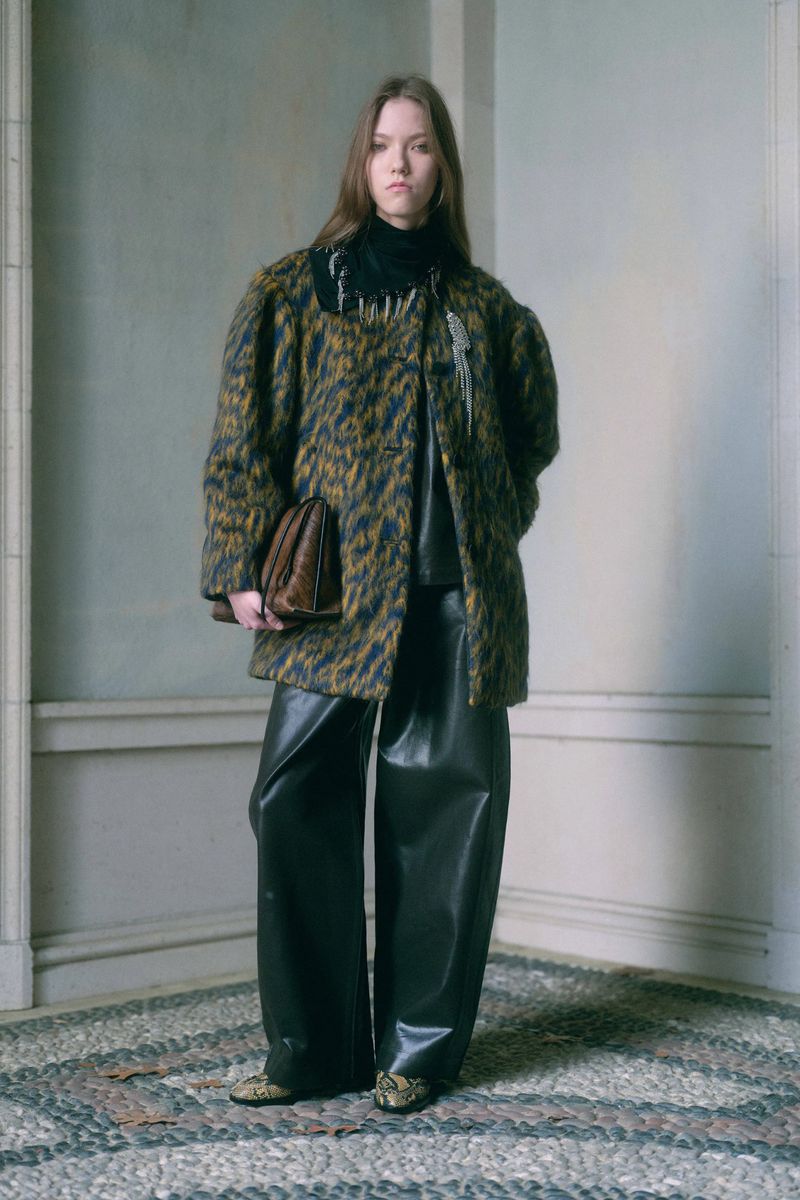 A young woman models a patterned coat, wide-leg leather pants, and a brown bag in a full shot indoors, under soft natural light.
