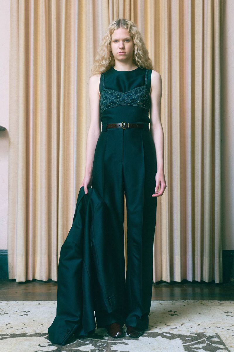 Fashion shot: a fair-skinned female model with curly blonde hair, wearing a sleeveless green top and matching pants. She is holding a jacket.