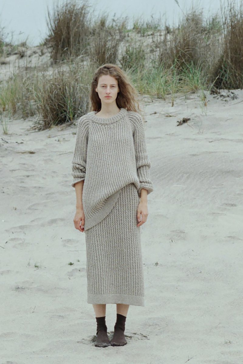 A young adult woman stands on a sandy beach wearing a knitted sweater and skirt, with beach grasses and a cloudy sky in the background.