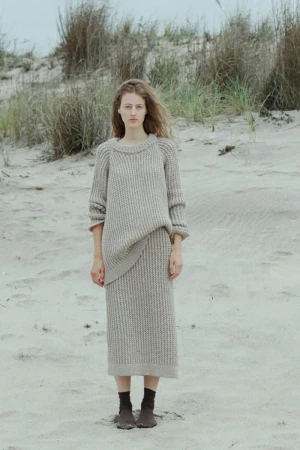 A young adult woman stands on a sandy beach wearing a knitted sweater and skirt, with beach grasses and a cloudy sky in the background.