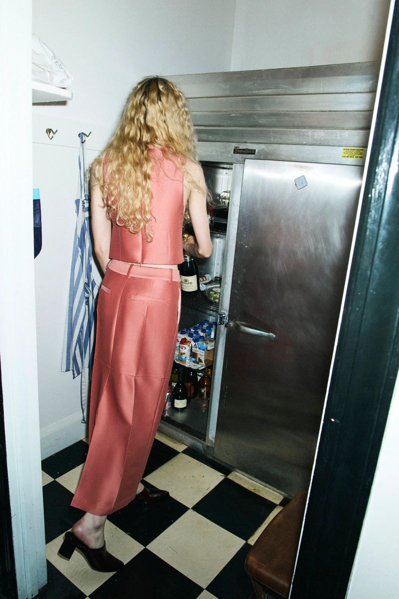 Woman in rose-colored outfit reaches into stainless steel refrigerator in a kitchen with black and white checkered floor and white walls.