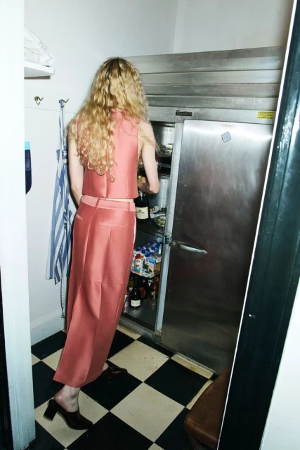 Woman in rose-colored outfit reaches into stainless steel refrigerator in a kitchen with black and white checkered floor and white walls.