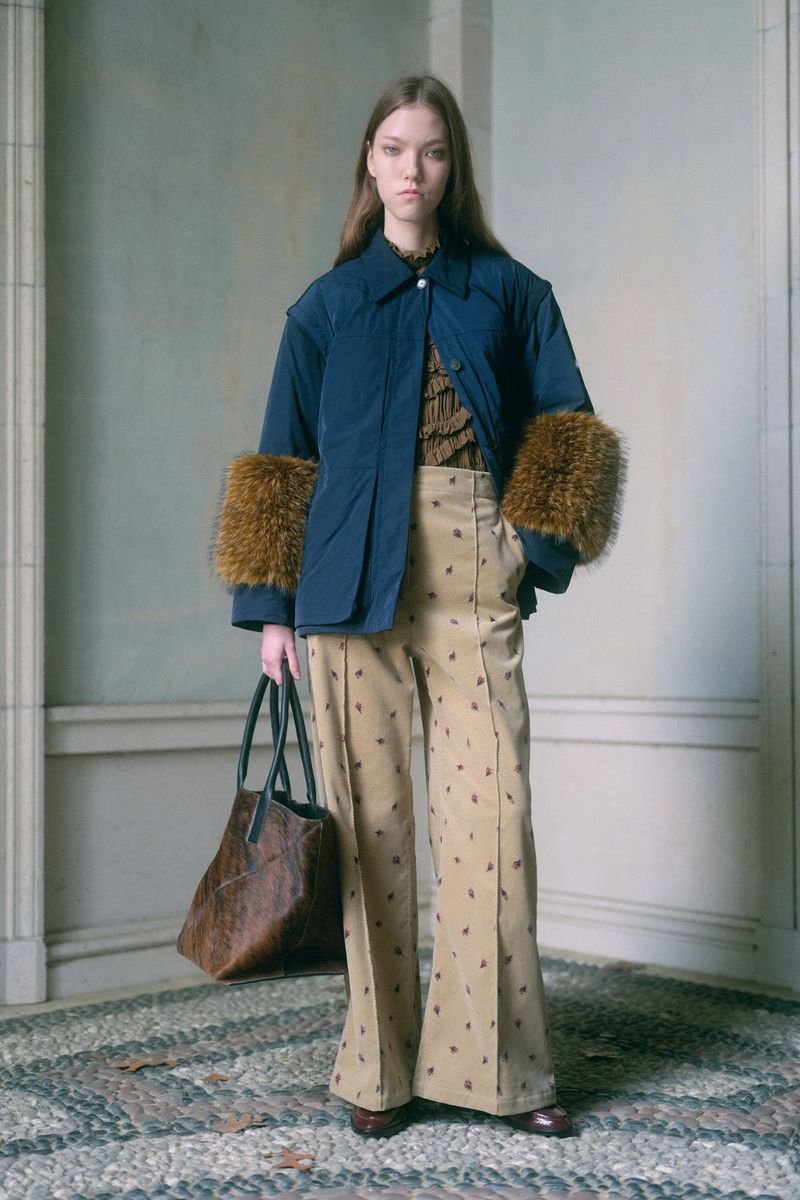 Fashion model poses in a blue jacket with fur sleeves and patterned tan pants, holding a brown leather tote bag indoors.