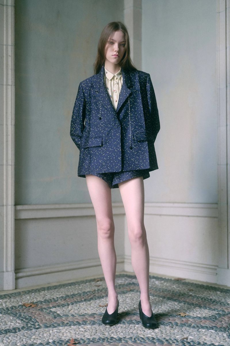 Fashion portrait of a model in a patterned blue blazer and shorts with black flats, posing indoors against a neutral wall with a stone floor.