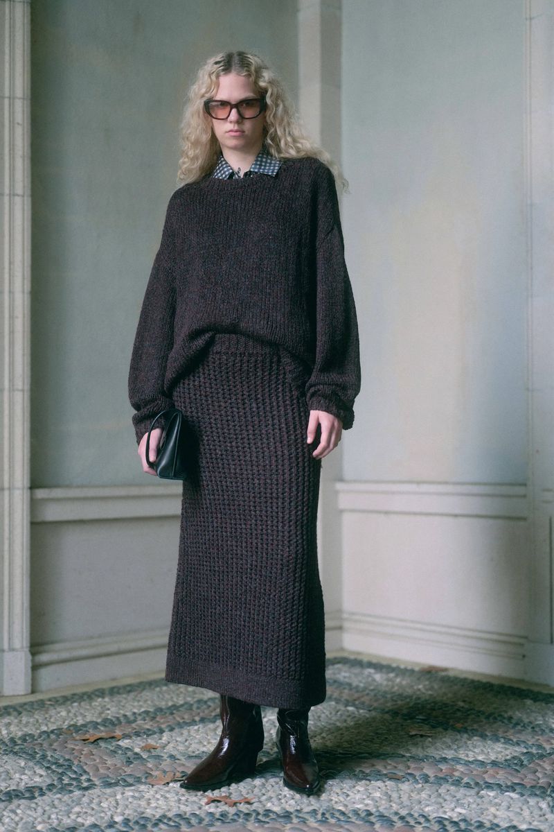 A blonde model poses in a brown knit sweater and matching skirt, accessorized with a black clutch and brown leather boots, standing indoors.