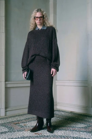 A blonde model poses in a brown knit sweater and matching skirt, accessorized with a black clutch and brown leather boots, standing indoors.