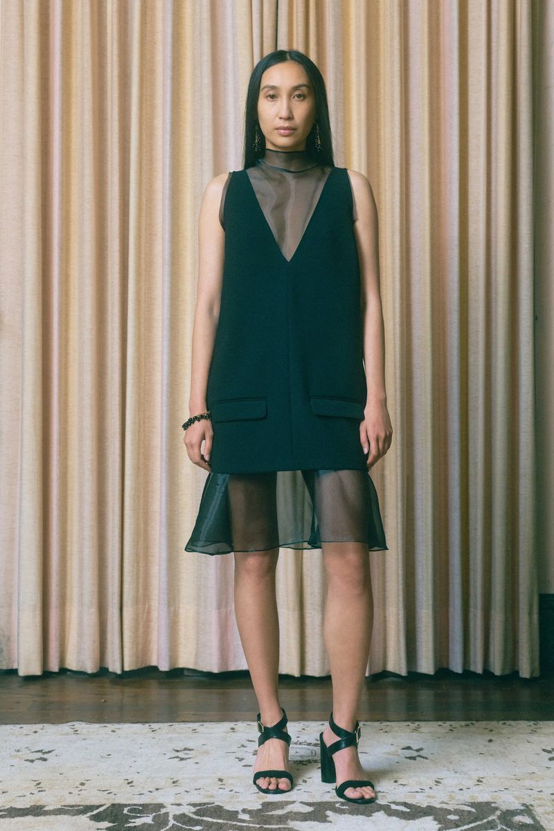 Adult female model in a sleeveless black dress and black high-heeled sandals poses in a photo studio with a beige curtain backdrop.