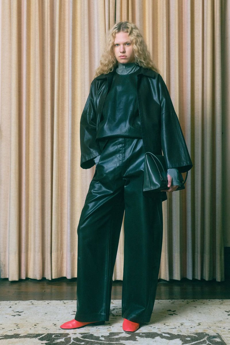 A woman with blonde wavy hair poses in a shiny black top, jacket, and pants with red shoes in front of a beige curtain backdrop.