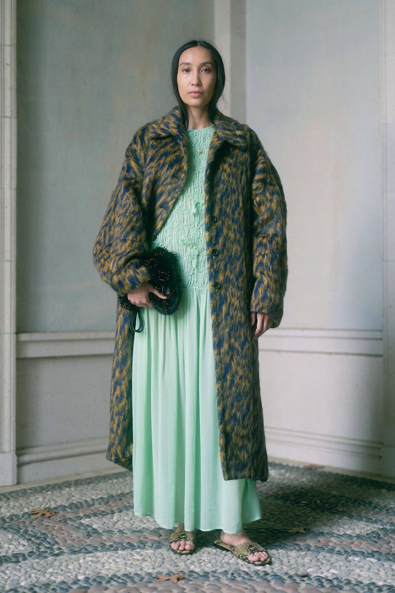 A woman poses in a blue and brown patterned coat over a green dress, holding a black purse, on a cobblestone path in soft light.