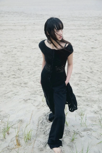 A slender Asian woman stands barefoot on a sandy beach, wearing a black knit dress over black pants, looking pensively off to the side.