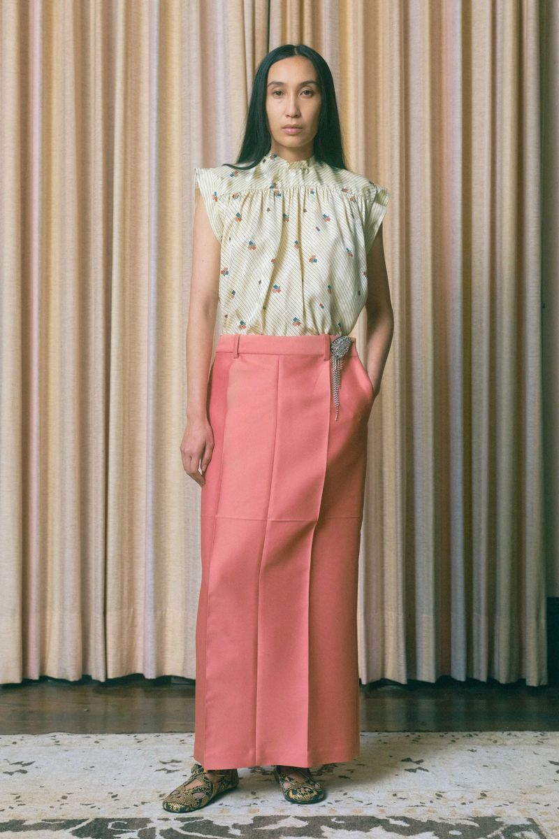 Full-length studio shot of a woman wearing a patterned, high-necked top and long pink skirt against a beige curtain backdrop.