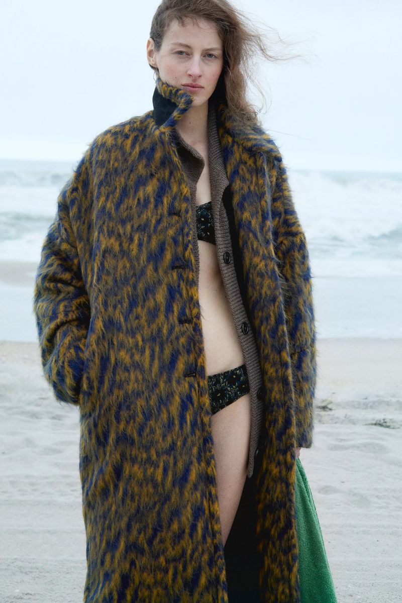 A woman in a bikini and patterned coat stands on a sandy beach with a neutral-toned ocean in the background under an overcast sky.