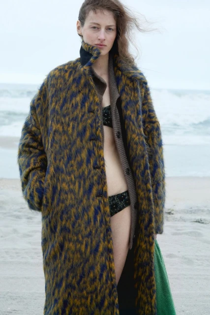 A woman in a bikini and patterned coat stands on a sandy beach with a neutral-toned ocean in the background under an overcast sky.