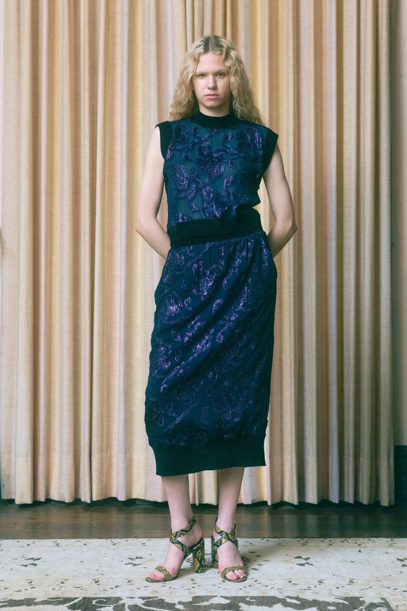 Woman posing in a blue floral-patterned knit dress and heeled sandals against a gold curtain backdrop in a full-length studio shot.