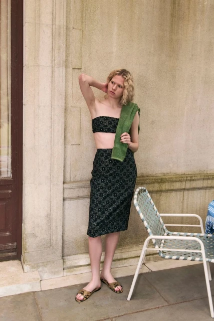 Woman in patterned two-piece set poses outdoors. She wears slides and holds a green towel, standing next to metal frame chairs.
