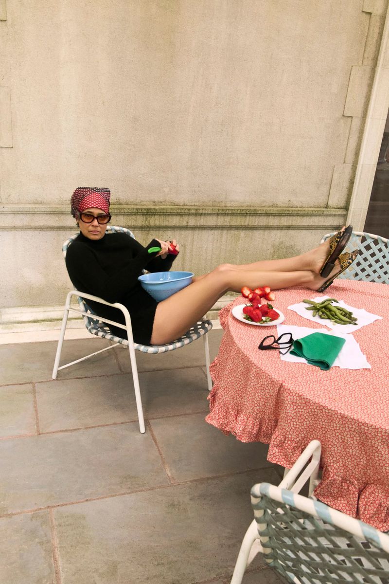 Woman in a black bodysuit sits in a chair with feet up on a table with a red patterned tablecloth and a bowl of fruit.