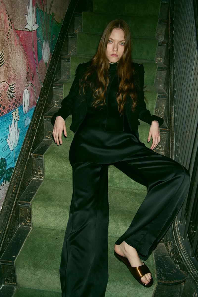 Brunette woman in black jacket and satin pants poses on green carpeted stairs with patterned wallpaper, creating a chic fashion image.