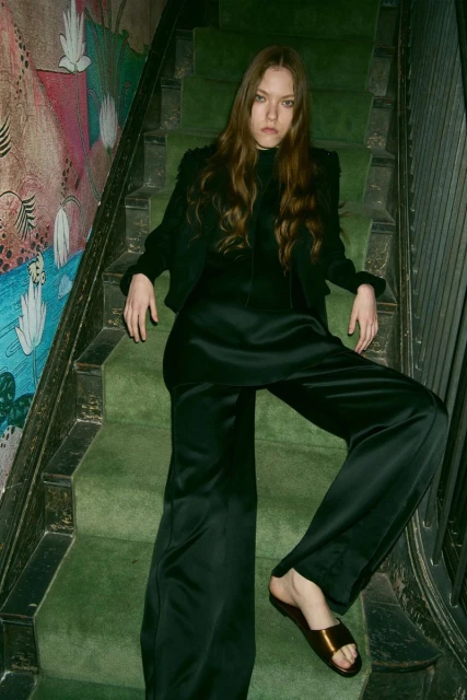 Brunette woman in black jacket and satin pants poses on green carpeted stairs with patterned wallpaper, creating a chic fashion image.