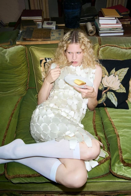 Woman with blonde curly hair lying on a green velvet couch while eating a dessert off a white plate, wearing a white dress and white socks.