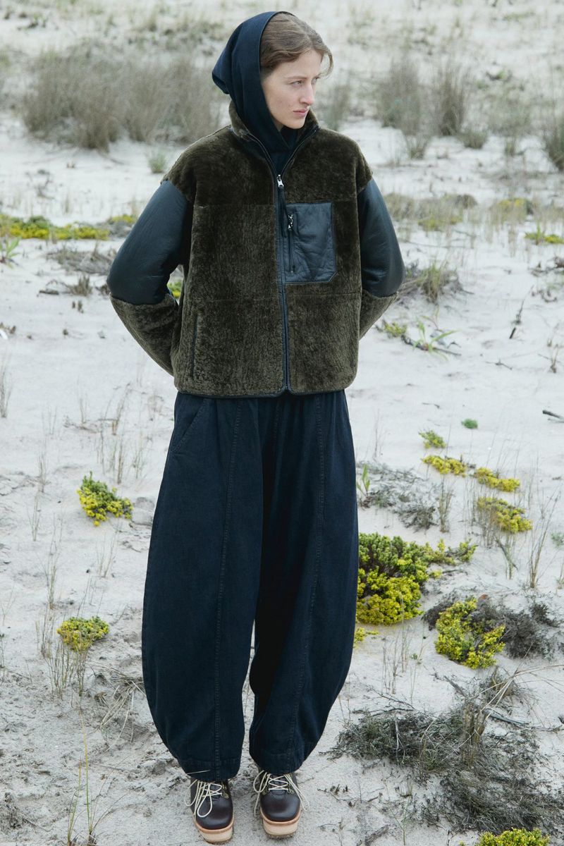 A young woman poses in a green fleece jacket and wide-legged pants on a sandy dune with sparse, low-lying vegetation on a cloudy day.