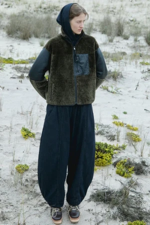 A young woman poses in a green fleece jacket and wide-legged pants on a sandy dune with sparse, low-lying vegetation on a cloudy day.