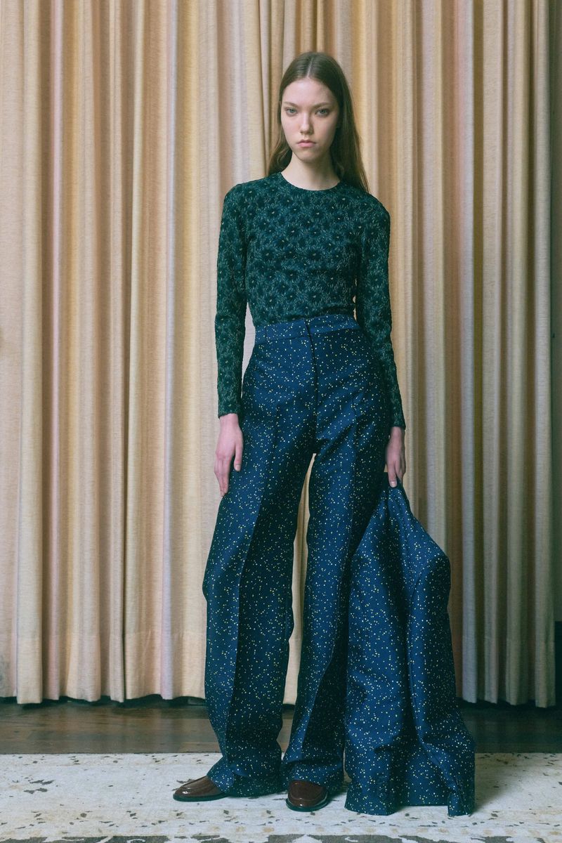 Full-length shot of a slender young woman in wide-leg patterned trousers and a floral sweater posing in front of a beige curtain.
