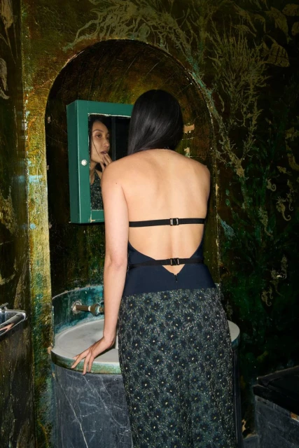 An Asian woman in a backless dress stands in a bathroom, facing a mirror with a green frame in a close shot.