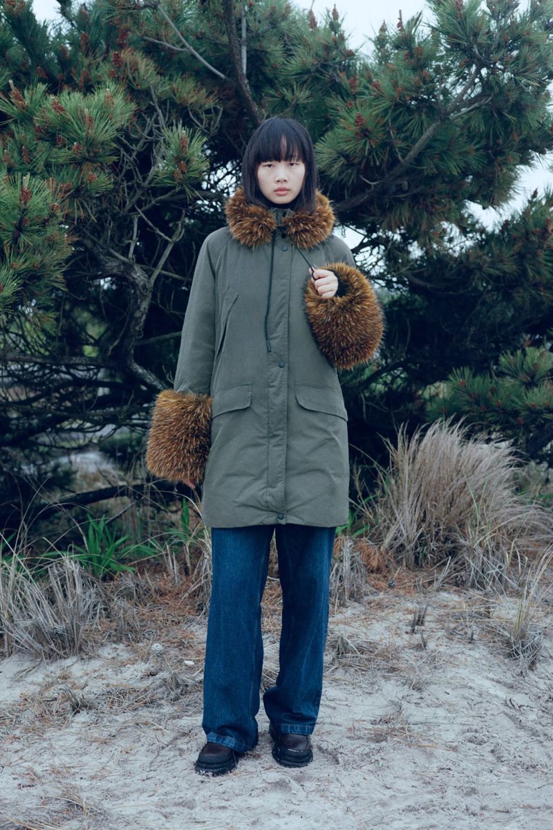 Adult woman wearing a parka with fur trim and wide-legged jeans stands outdoors near a pine tree and grassy terrain.