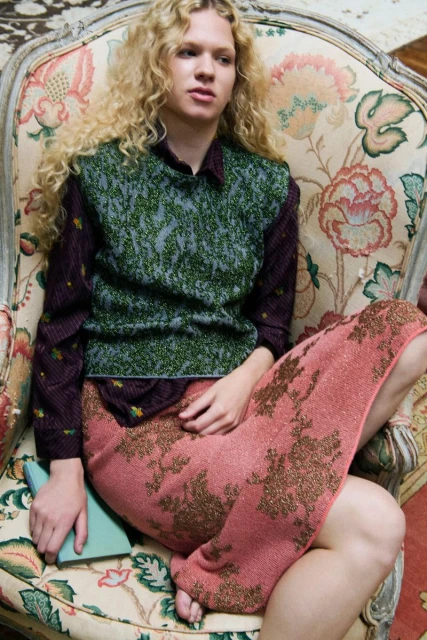 A young adult with long blonde curls sits on an antique chair, wearing a patterned sweater vest, long skirt, and holding a light blue notebook.