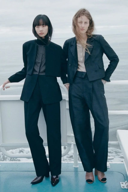 Two women in modern formalwear pose outdoors with a neutral backdrop, emphasizing sleek blazers, wide-leg pants, and contemporary style.