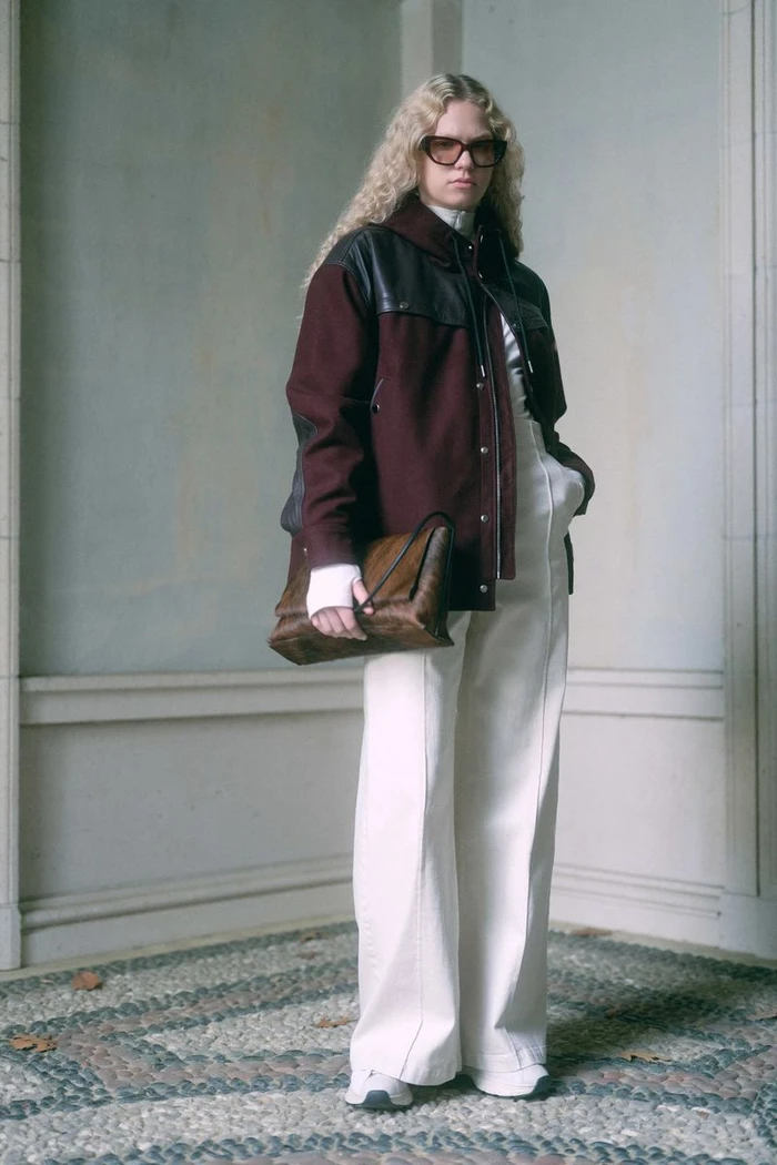 Model with long blonde curly hair wearing dark sunglasses, a burgundy jacket, and white trousers holding a brown leather bag.