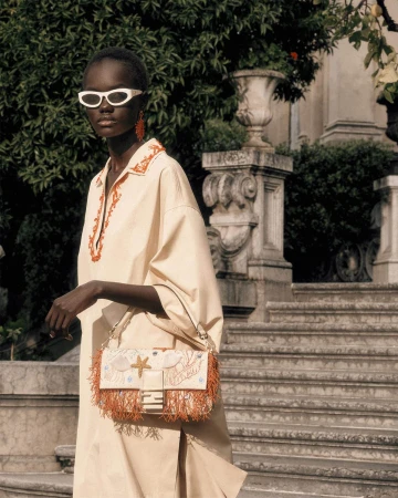 Model Achol Kuir wearing white sunglasses and a cream tunic dress, holding an embellished Fendi handbag on stone steps.