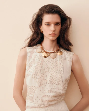 Model wearing a patterned white top and statement shell necklace against a warm neutral studio background.
