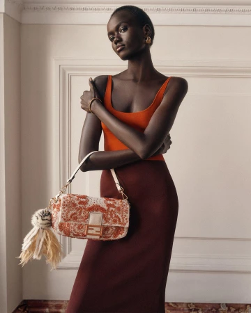 Model Achol Kuir wearing an orange top and burgundy skirt holding a patterned Fendi handbag indoors against panelled walls