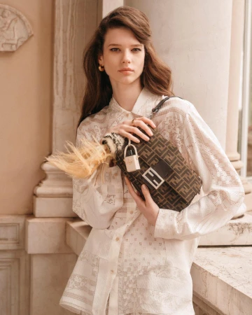 Model holding a brown monogram shoulder bag wearing a patterned white shirt outdoors near stone architectural columns