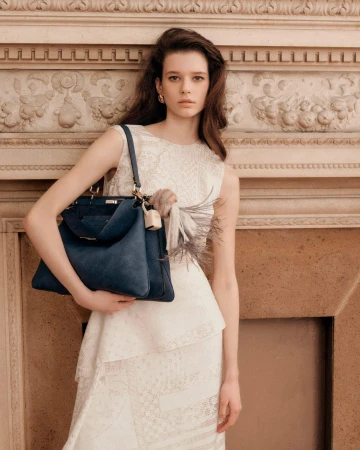 Model holding a dark blue handbag wearing an off-white lace dress standing in front of an ornate stone fireplace.