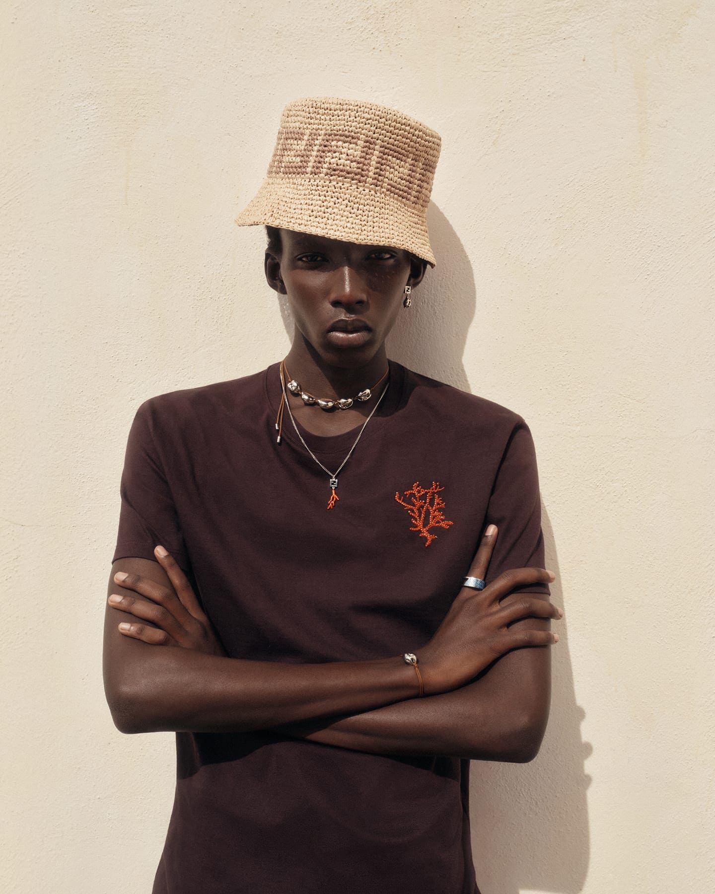 Model Achol Kuir wearing a straw bucket hat and dark brown t-shirt with arms crossed against a plain wall.