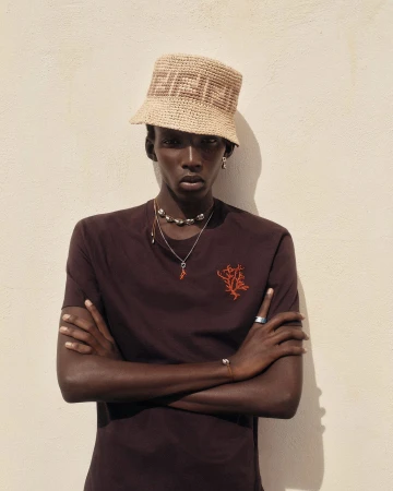 Model Achol Kuir wearing a straw bucket hat and dark brown t-shirt with arms crossed against a plain wall.