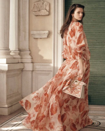 Model wearing a long floral patterned dress holding a white handbag in front of classical architectural columns.