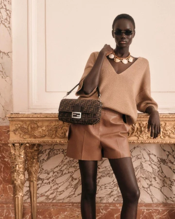 Model Achol Kuir wearing brown shorts and tan sweater holding a Fendi monogram handbag indoors