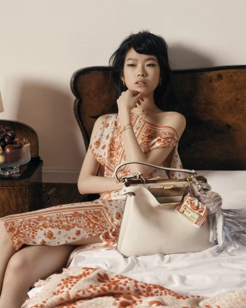 Model wearing a patterned orange and white dress seated on a bed with a white leather handbag in the foreground