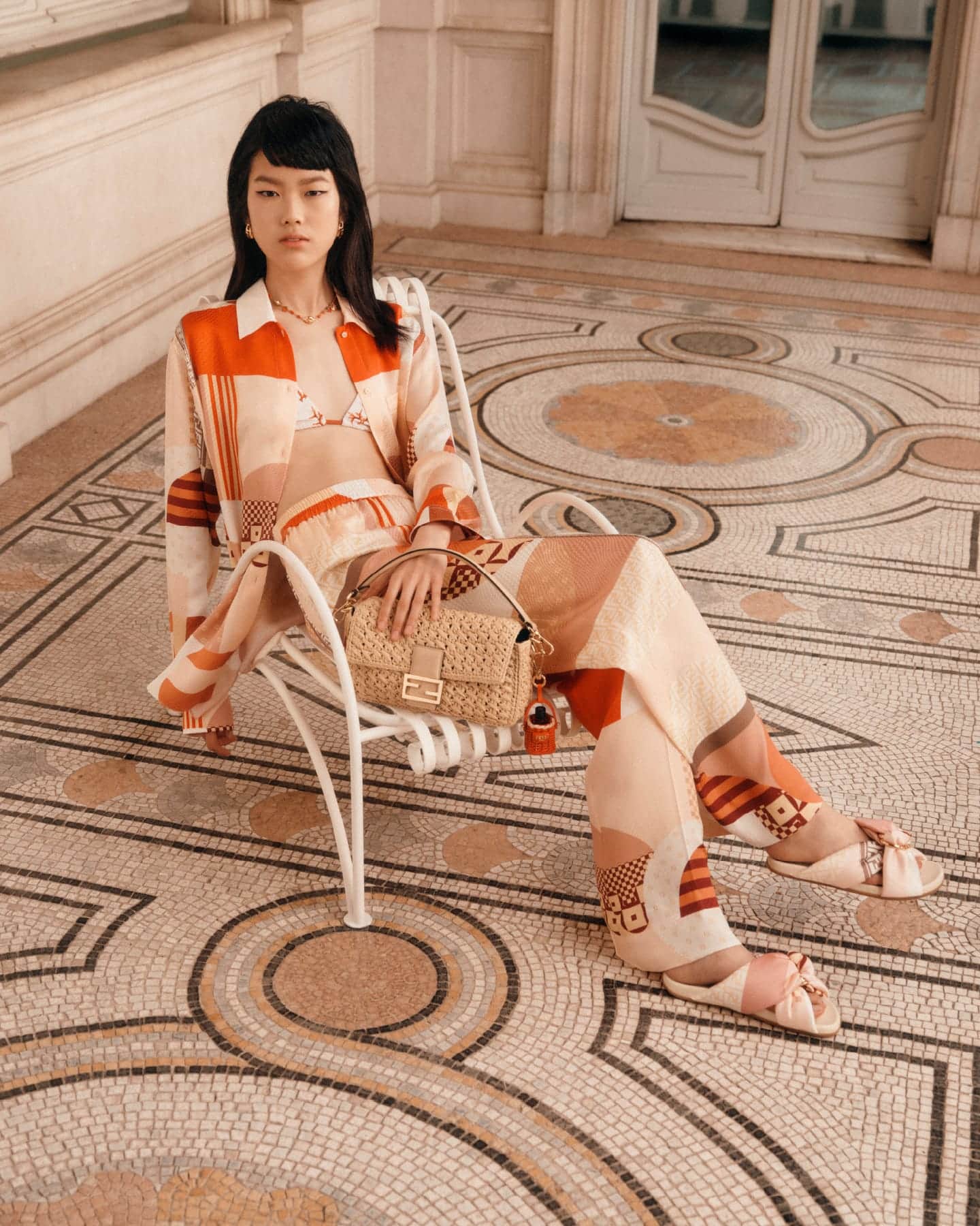 Model seated on a white metal chair wearing a patterned orange shirt and trousers holding a woven handbag