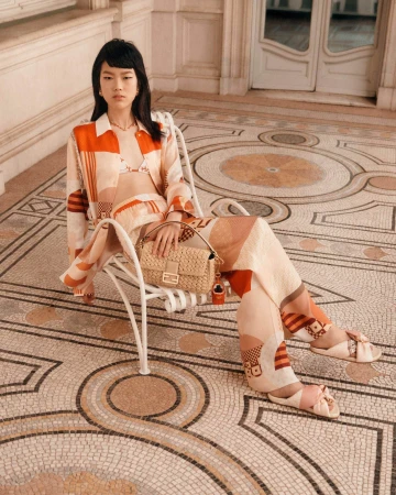 Model seated on a white metal chair wearing a patterned orange shirt and trousers holding a woven handbag