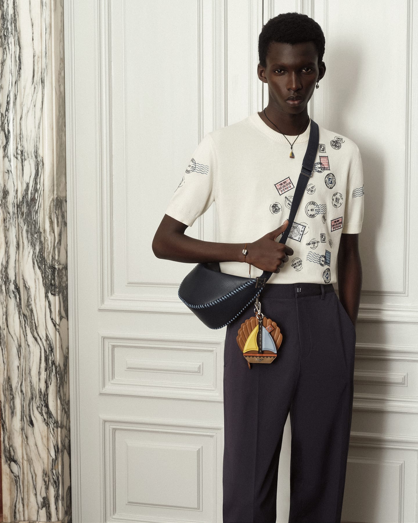 Model Achol Kuir wearing a patterned white t-shirt and dark trousers while carrying a black cross-body bag indoors.