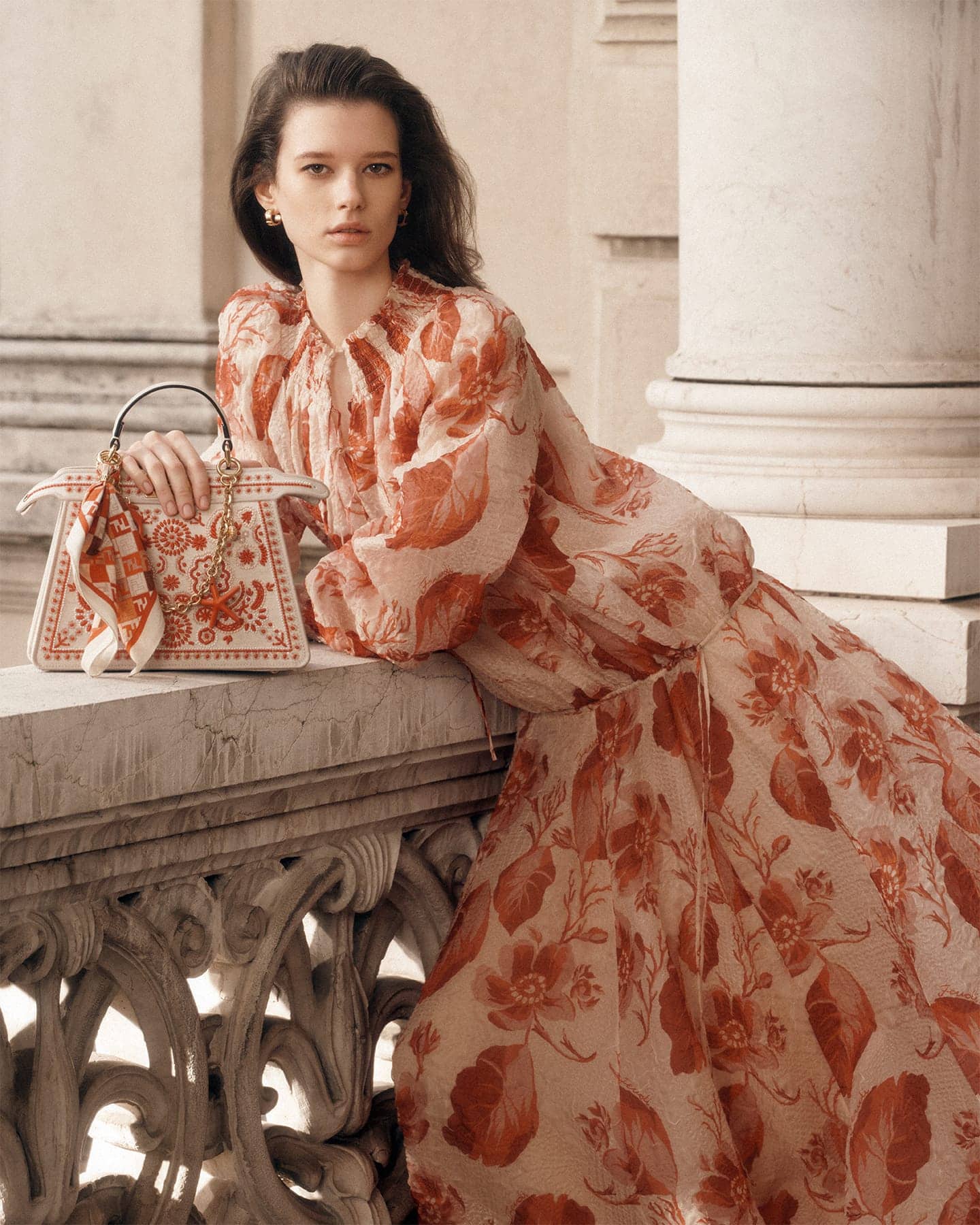 Model in a patterned red and white dress holding a patterned handbag, leaning against an ornate stone balcony.