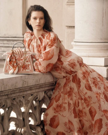 Model in a patterned red and white dress holding a patterned handbag, leaning against an ornate stone balcony.
