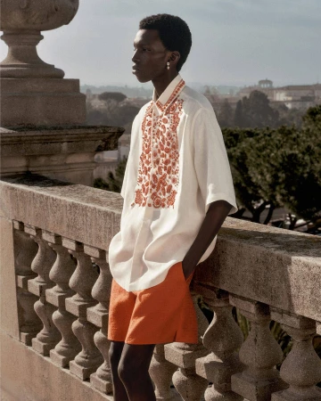 Model leaning on a stone balustrade wearing an embroidered white short-sleeve shirt and orange shorts