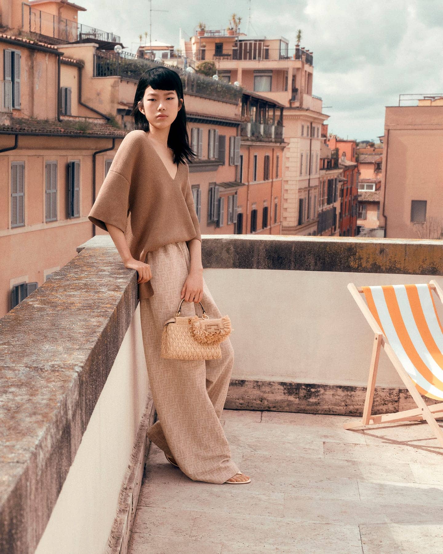 Model leaning on a stone balcony holding a woven handbag against a city rooftop background.