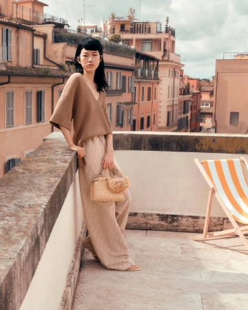 Model leaning on a stone balcony holding a woven handbag against a city rooftop background.