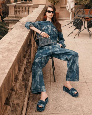A model in a blue denim floral patterned outfit seated on a terrace leaning against a stone balustrade holding a handbag